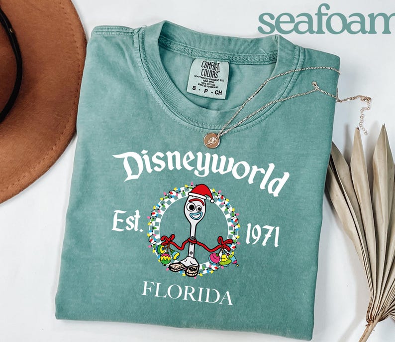 May include: Seafoam green t-shirt with the text "Disneyworld Est. 1971 Florida". The shirt features a cartoon fork character wearing a Santa hat and holding Christmas ornaments. A gold necklace is draped across the shirt.