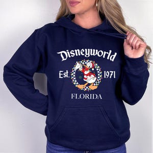 Pu&ograve; includere: Felpa con cappuccio blu navy con la scritta "Disneyworld Est. 1971 FLORIDA". La grafica presenta Paperino con un cappello da Babbo Natale e una giacca rossa, circondato da una ghirlanda di luci e un bordo a scacchi.