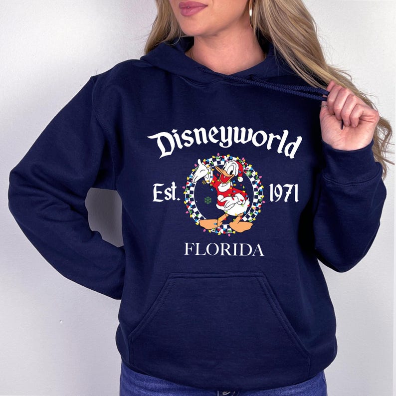 Pu&ograve; includere: Felpa con cappuccio blu navy con la scritta "Disneyworld Est. 1971 FLORIDA". La grafica presenta Paperino con un cappello da Babbo Natale e una giacca rossa, circondato da una ghirlanda di luci e un bordo a scacchi.