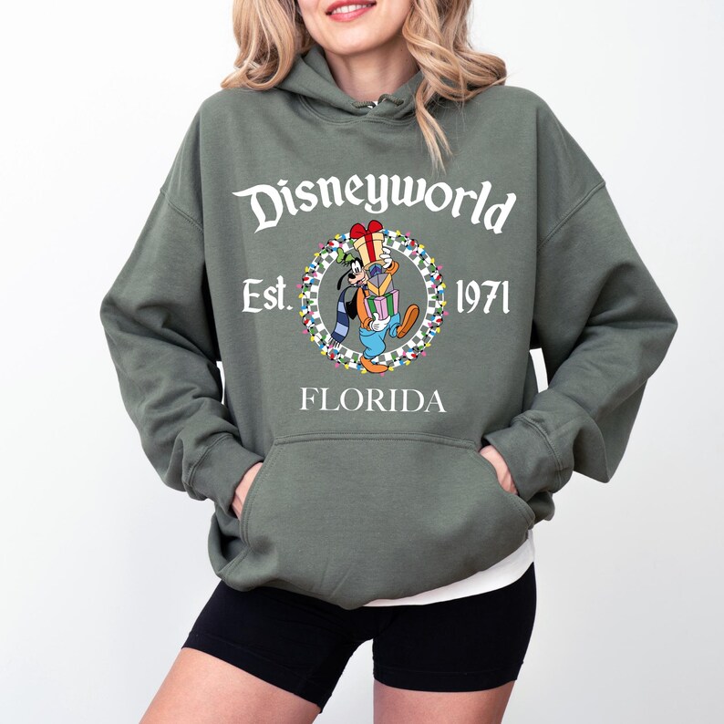 Pu&ograve; includere: Una felpa con cappuccio verde oliva con una tasca frontale. La felpa presenta la scritta "Disneyworld" sopra una grafica di Pippo che tiene dei regali, con "Est. 1971 FLORIDA" sotto. La felpa &egrave; indossata sopra una maglietta bianca e pantaloncini neri.