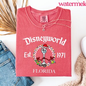 May include: A red t-shirt with the text "Disneyworld Est. 1971 FLORIDA" in white. The shirt features a cartoon character wearing a Santa hat. A silver necklace is on the shirt. The word "watermelon" is in the top right corner.