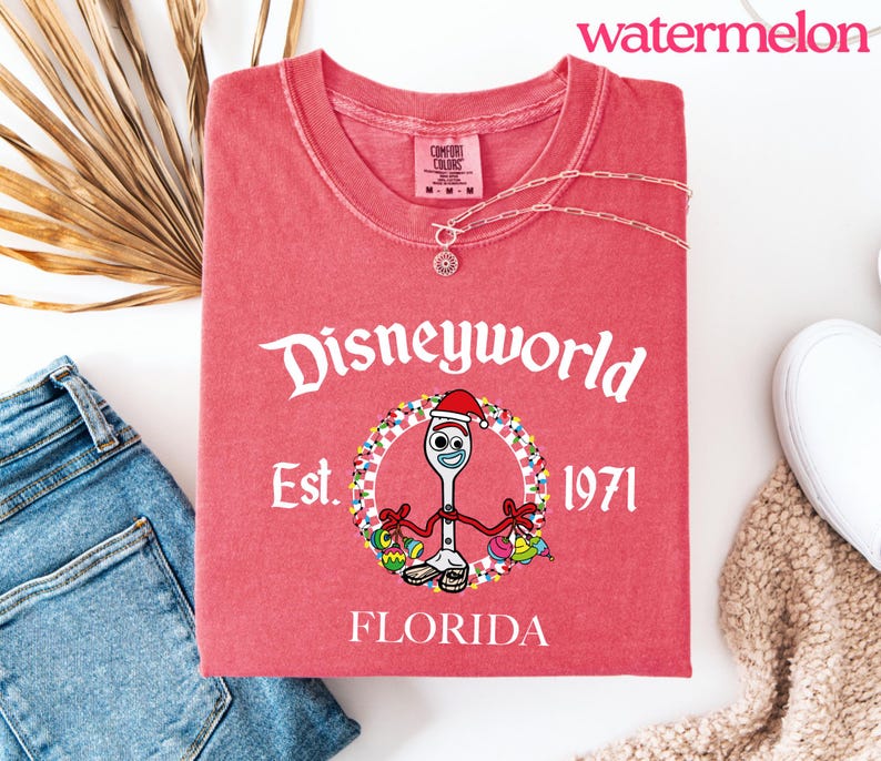 May include: A red t-shirt with the text "Disneyworld Est. 1971 FLORIDA" in white. The shirt features a cartoon character wearing a Santa hat. A silver necklace is on the shirt. The word "watermelon" is in the top right corner.