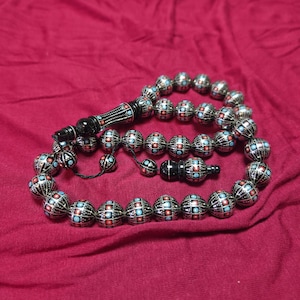 Peut inclure: Un chapelet composé de perles noires sphériques ornées de motifs linéaires argentés complexes et de petites incrustations circulaires rouges et bleu clair. Le chapelet comprend une perle noire cylindrique plus longue et un pompon.