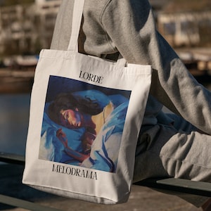 Puede incluir: Bolso tote de color crema con la palabra "LORDE" encima de una pintura de una persona en la cama, con la palabra "MELODRAMA" debajo. La obra de arte est&aacute; en tonos azules, blancos y melocot&oacute;n. El bolso tiene dos correas largas.