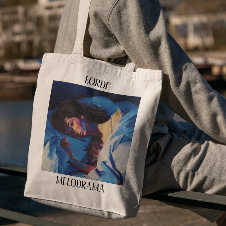 Puede incluir: Bolso tote de color crema con la palabra "LORDE" encima de una pintura de una persona en la cama, con la palabra "MELODRAMA" debajo. La obra de arte est&aacute; en tonos azules, blancos y melocot&oacute;n. El bolso tiene dos correas largas.