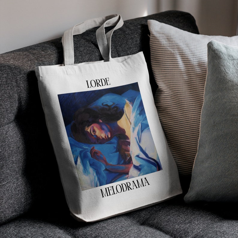 Puede incluir: Bolso tote blanco con la portada del &aacute;lbum "Melodrama" de Lorde. La obra de arte representa a una persona en una cama con s&aacute;banas azules. La palabra "LORDE" est&aacute; impresa encima de la imagen, y "MELODRAMA" est&aacute; impresa debajo.