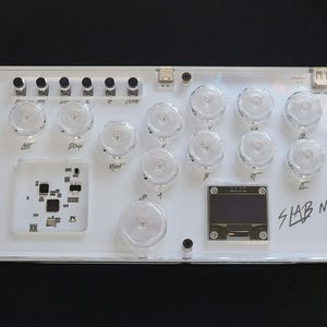Könnte beinhalten: Ein weißer Musikcontroller aus Kunststoff mit einem klaren Display und 12 runden Knöpfen. Der Text "SLAB MINI V1.3" ist auf dem Controller aufgedruckt.