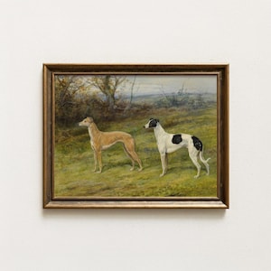 May include: Framed vintage painting of two greyhounds in a field. One dog is tan, the other is white with black spots. The artwork is set in a gold-toned frame, with a landscape background of trees and a cloudy sky.