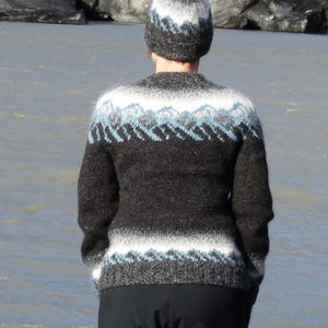 Peut inclure: Un pull en tricot à manches longues et un bonnet assorti présentant une base gris anthracite foncé avec des bandes horizontales de motifs de montagne blancs et bleu clair sur la poitrine, les épaules et l'ourlet du pull et du bonnet.