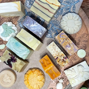 May include: Assortment of handmade soaps in various shapes and colors, including bars with swirls, floral designs, and honeycomb textures. Soaps are arranged on a wooden tray with a floral patterned cloth.