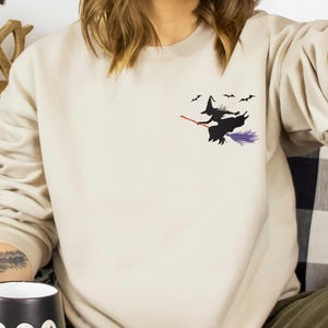 May include: A beige sweatshirt featuring a small graphic of a witch on a broomstick with bats. The witch is black with a purple skirt and red broom. The mug in the person's hand has the word "Boo" and ghost graphics.