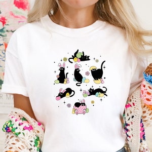 May include: White t-shirt with a black cat wearing bunny ears and holding Easter eggs. The cat is surrounded by colorful Easter eggs and flowers.
