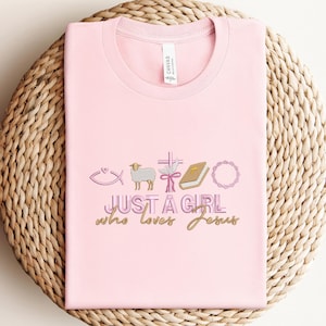 May include: Pink t-shirt with a graphic design of religious symbols and the text "JUST A GIRL who loves Jesus".