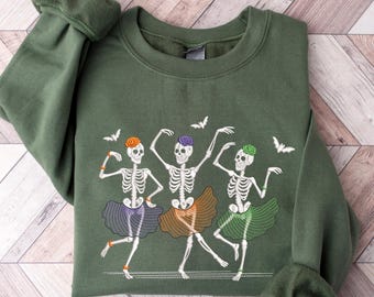 May include: An olive green sweatshirt featuring a whimsical design of three dancing skeletons in tutus. The skeletons have orange, purple, and green accents, with bats flying above them. The sweatshirt is folded on a light wood-grain surface.