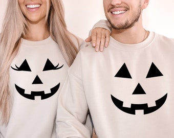 May include: A couple wearing matching white sweatshirts with black jack-o'-lantern faces printed on the front. The woman's sweatshirt has eyelashes on the jack-o'-lantern face.