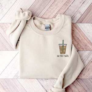 May include: A beige sweatshirt with a graphic of a brown iced coffee drink with a green straw and the text "on the rocks" printed on the front.