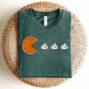 May include: Green t-shirt featuring a pie slice design with three dollops of whipped cream. The pie slice is orange with a brown crust, and the whipped cream is white. The shirt is made of a soft material and is displayed on a woven surface.
