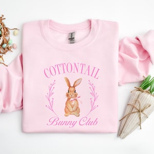May include: Pink sweatshirt with a watercolor illustration of a brown bunny with a pink bow. The bunny is surrounded by pink floral sprigs and the text "COTTONTAIL Bunny Club".