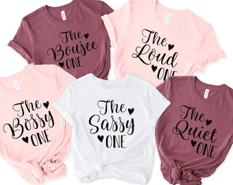 May include: Five pink, white, and maroon t-shirts with black text that reads "The Boujee One", "The Bossy One", "The Sassy One", "The Loud One", and "The Quiet One".