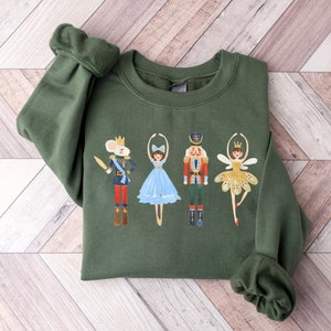 May include: A green sweatshirt with a festive design featuring a mouse, two ballerinas, and a nutcracker. The ballerinas are wearing blue and gold tutus.