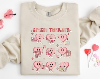 May include: A beige sweatshirt with the text "Speech Therapy" printed in pink and red letters. The design features nine pink hearts with different speech therapy tools and activities.