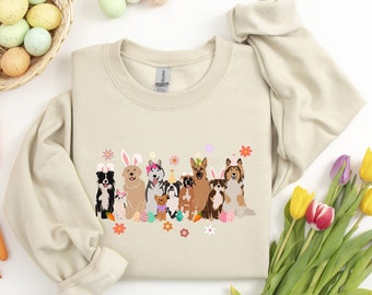 May include: A beige sweatshirt with a graphic of dogs wearing bunny ears and other Easter-themed accessories. The dogs are surrounded by flowers and Easter eggs.