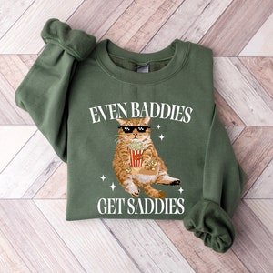 May include: Green sweatshirt with a cartoon cat wearing sunglasses and holding a bucket of popcorn. The text on the sweatshirt reads "Even Baddies Get Saddies".