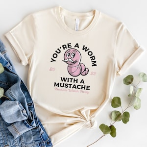 May include: A beige t-shirt with a graphic of a pink worm with a mustache and the text "You're a worm with a mustache Reunion Watch Party 2023".