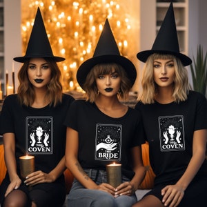 May include: Three women wearing black witch hats and black t-shirts with white tarot card designs. The shirts say "The Coven", "The Bride", and "The Coven".