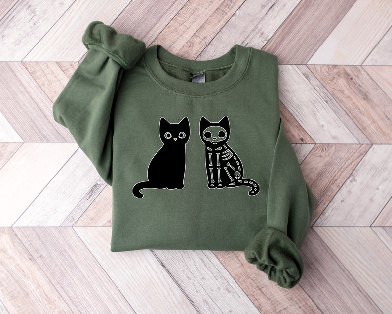 May include: A green sweatshirt with a black cat and a skeleton cat graphic. The cats are facing each other.