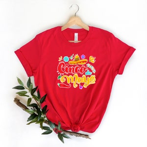 May include: Red t-shirt with a colorful graphic design that says "Cinco de Mayo" with a sombrero and flowers.