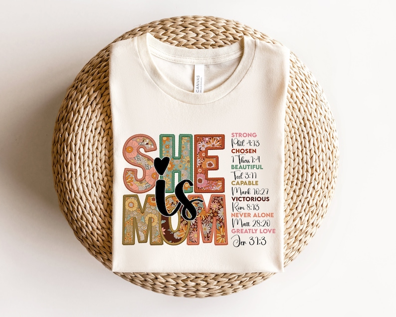 May include: A white t-shirt with a floral design that says "She is Mom" with bible verses listed next to each letter. The verses are: Strong Phil 4:13, Chosen 1 Thess 1:4, Beautiful Tal 3:11, Capable Mark 10:27, Victorious Rom 8:13, Never Alone Matt 28:20, Greatly Love Jer 31:3.