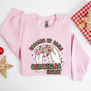 Puede incluir: Sudadera rosa de Navidad con un gr&aacute;fico de manos enrollando un porro con el texto "Rolling Up Some Christmas Spirit".
