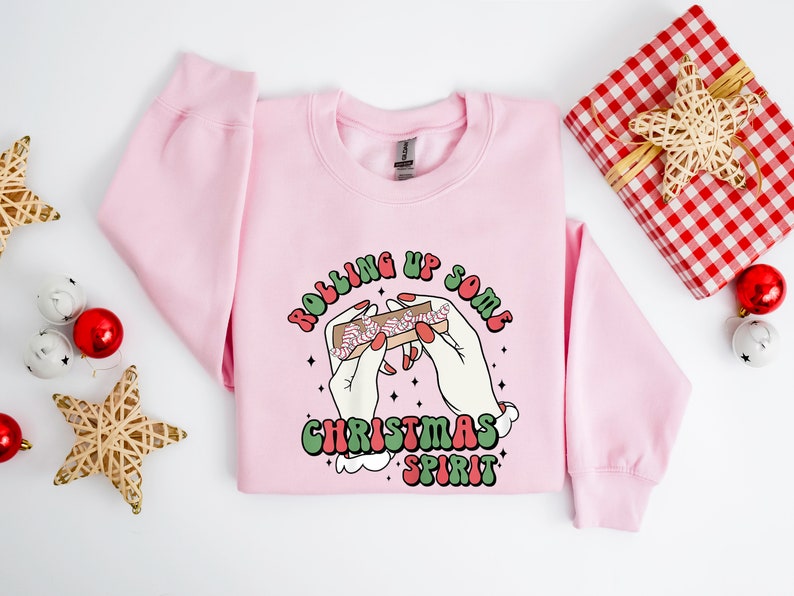 Puede incluir: Sudadera rosa de Navidad con un gr&aacute;fico de manos enrollando un porro con el texto "Rolling Up Some Christmas Spirit".