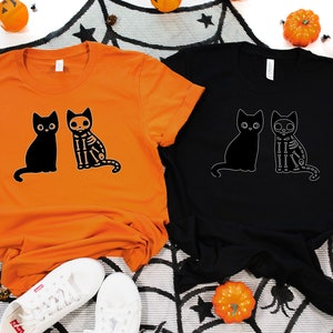 May include: Two t-shirts with a black cat and a skeleton cat design. The t-shirts are orange and black.