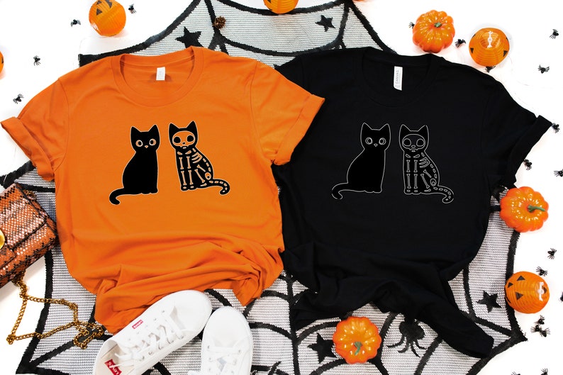 May include: Two t-shirts with a black cat and a skeleton cat design. The t-shirts are orange and black.