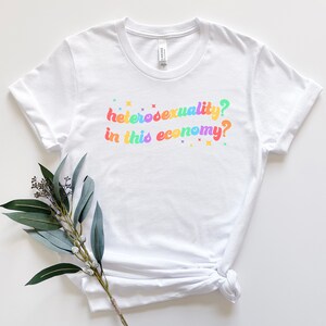 Pu&ograve; includere: Maglietta bianca con la scritta "eterosessualit&agrave;? in questa economia?" stampata in colori arcobaleno.