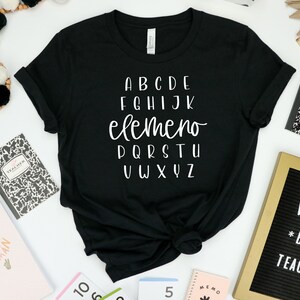 以下が含まれることがあります： 白い文字で「ABCDE FGHIJK elemeno PQRSTU VWXYZ」と書かれた黒いTシャツ。Tシャツは、ノート、鉛筆、そして「WORLDS *BEST* TEACHER」と書かれた文字盤など、他の学用品と一緒に白い表面に置かれています。