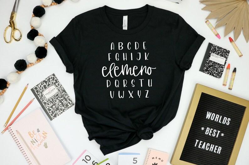 以下が含まれることがあります： 白い文字で「ABCDE FGHIJK elemeno PQRSTU VWXYZ」と書かれた黒いTシャツ。Tシャツは、ノート、鉛筆、そして「WORLDS *BEST* TEACHER」と書かれた文字盤など、他の学用品と一緒に白い表面に置かれています。