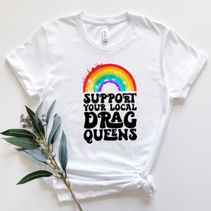 May include: White t-shirt with a rainbow graphic and the text "Support Your Local Drag Queens" in black.