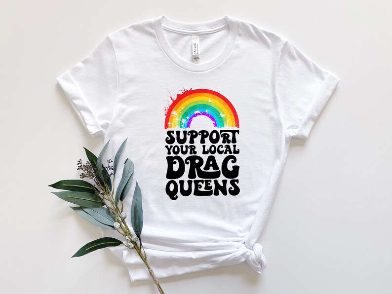 May include: White t-shirt with a rainbow graphic and the text "Support Your Local Drag Queens" in black.