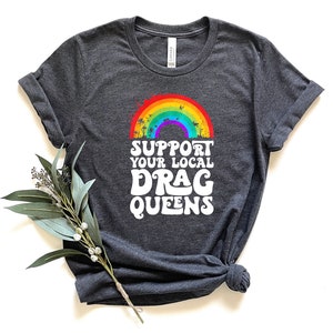 May include: A dark heather grey t-shirt with a rainbow and palm trees graphic. The text "Support Your Local Drag Queens" is printed in white below the graphic.
