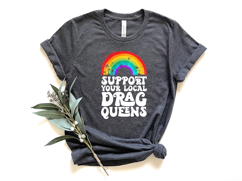 May include: A dark heather grey t-shirt with a rainbow and palm trees graphic. The text "Support Your Local Drag Queens" is printed in white below the graphic.
