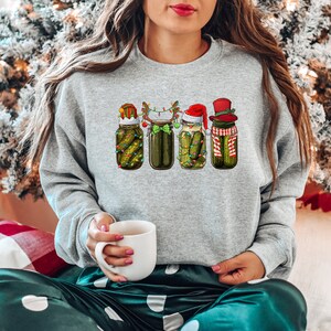 Pu&ograve; includere: Una felpa grigia con un design natalizio che presenta quattro barattoli di sottaceti con cappelli e accessori festivi. I barattoli sono decorati con luci e sottaceti verdi. La felpa &egrave; indossata da una persona seduta su una coperta a quadri.