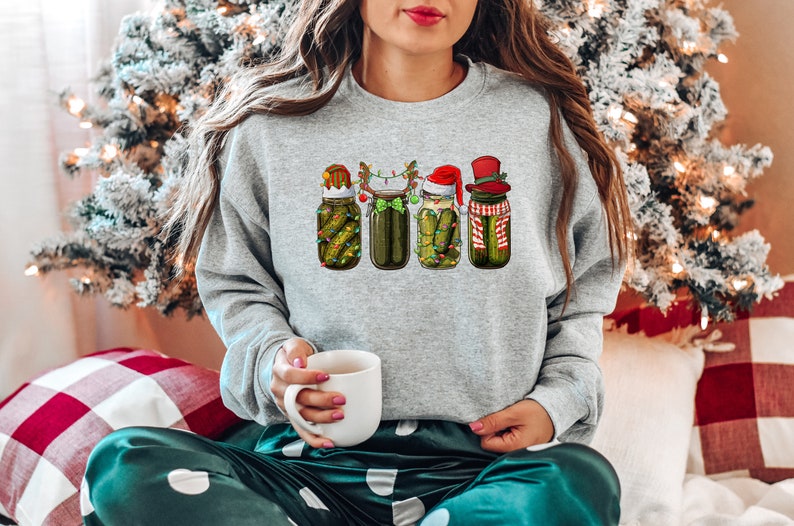 Pu&ograve; includere: Una felpa grigia con un design natalizio che presenta quattro barattoli di sottaceti con cappelli e accessori festivi. I barattoli sono decorati con luci e sottaceti verdi. La felpa &egrave; indossata da una persona seduta su una coperta a quadri.