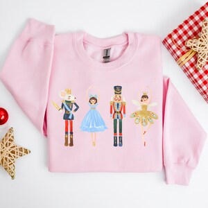 May include: Pink sweatshirt with a festive design featuring a mouse king, a ballerina, the Nutcracker, and a sugar plum fairy. The characters are all wearing colorful costumes and are standing in a line.
