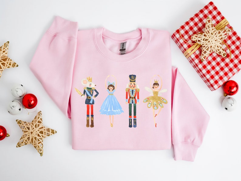May include: Pink sweatshirt with a festive design featuring a mouse king, a ballerina, the Nutcracker, and a sugar plum fairy. The characters are all wearing colorful costumes and are standing in a line.