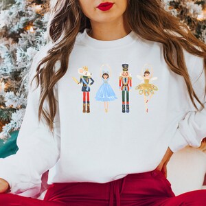 May include: A white sweatshirt with a festive design featuring a nutcracker, a ballerina, a mouse, and a crown. The design is in a whimsical style with bright colors.