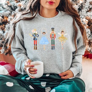May include: A gray sweatshirt with a festive design featuring a mouse, a ballerina, a nutcracker, and a fairy. The design is in a cartoon style with bright colors.