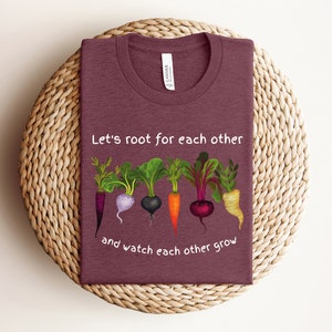 May include: A maroon t-shirt with the text "Let's root for each other and watch each other grow" printed in white. The text is surrounded by a graphic of five different root vegetables.